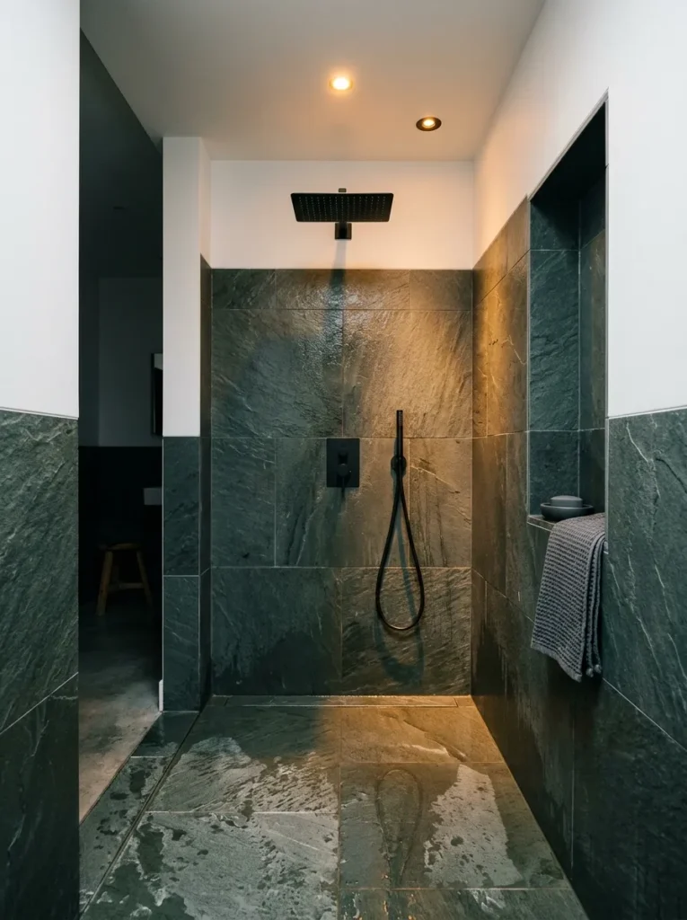 Moody wet room spa bathroom with matte black fixtures, dark slate floor tiles, wall-mounted rain head, and recessed amber lighting