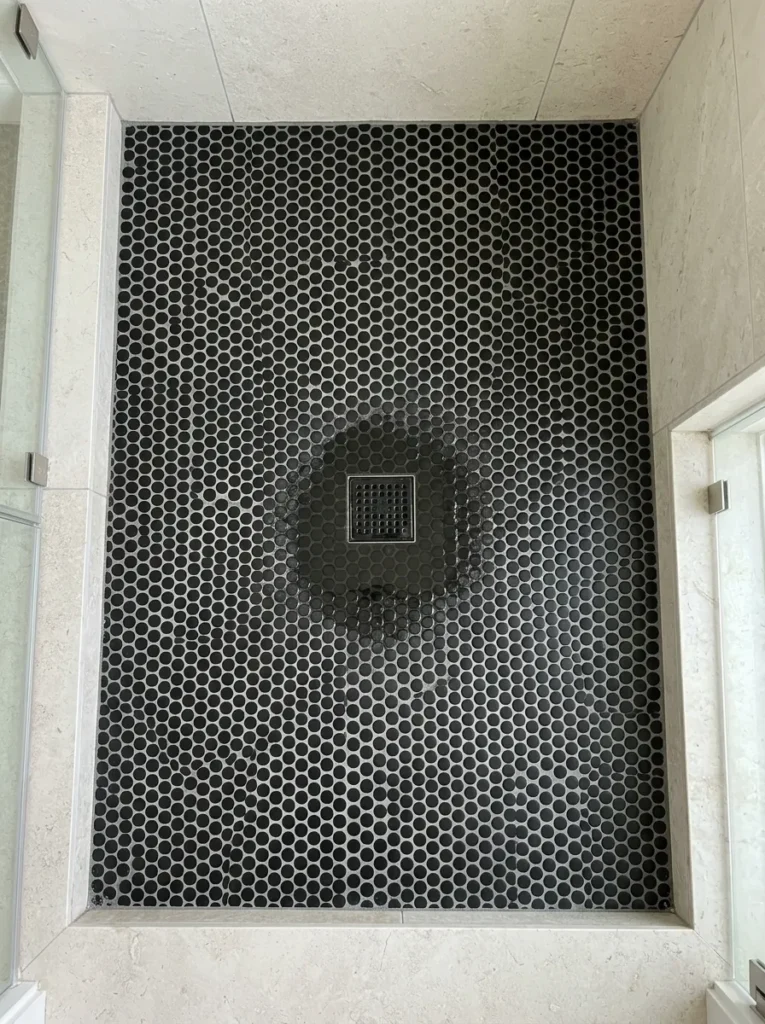 Matte black penny mosaic tiles covering a shower floor viewed from above, with grey grout lines and pale stone wall tiles surrounding