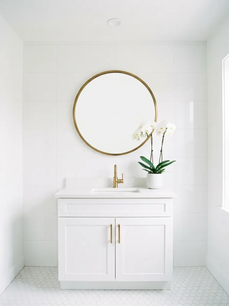 Round brushed brass mirror as the sole warm accent above a white shaker vanity in a fully white-tiled bathroom
