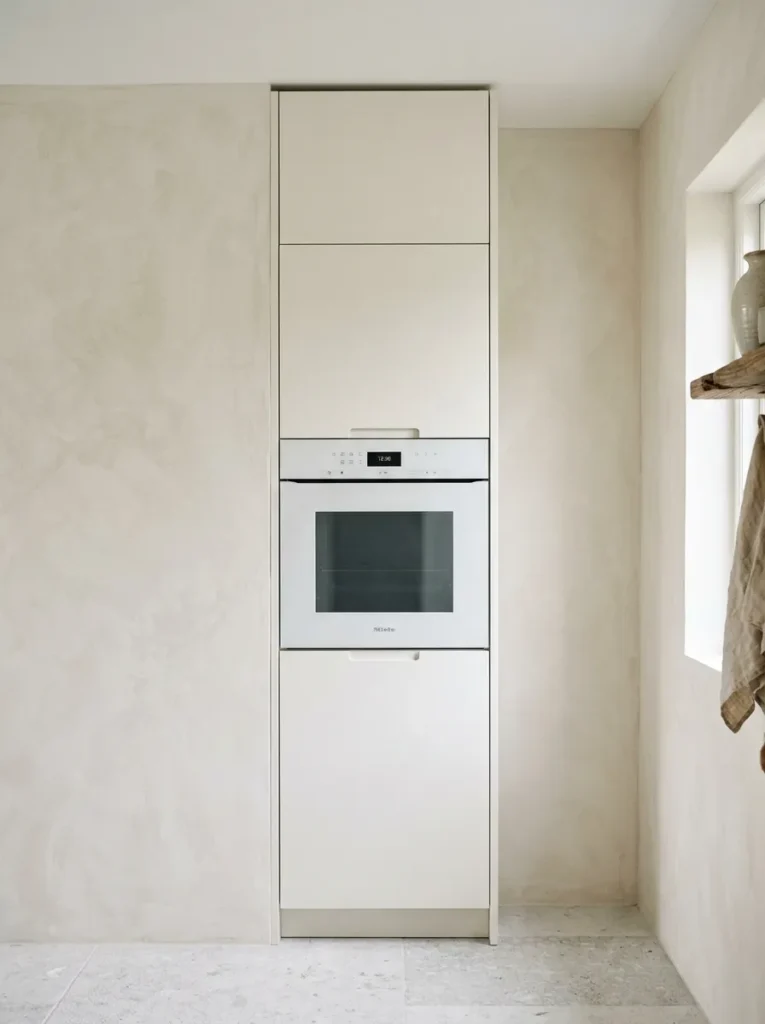 Floor-to-ceiling kitchen tower unit in warm white matte cabinetry with a flush-set built-in oven, no projecting handle, blending seamlessly into the vertical cabinet run
