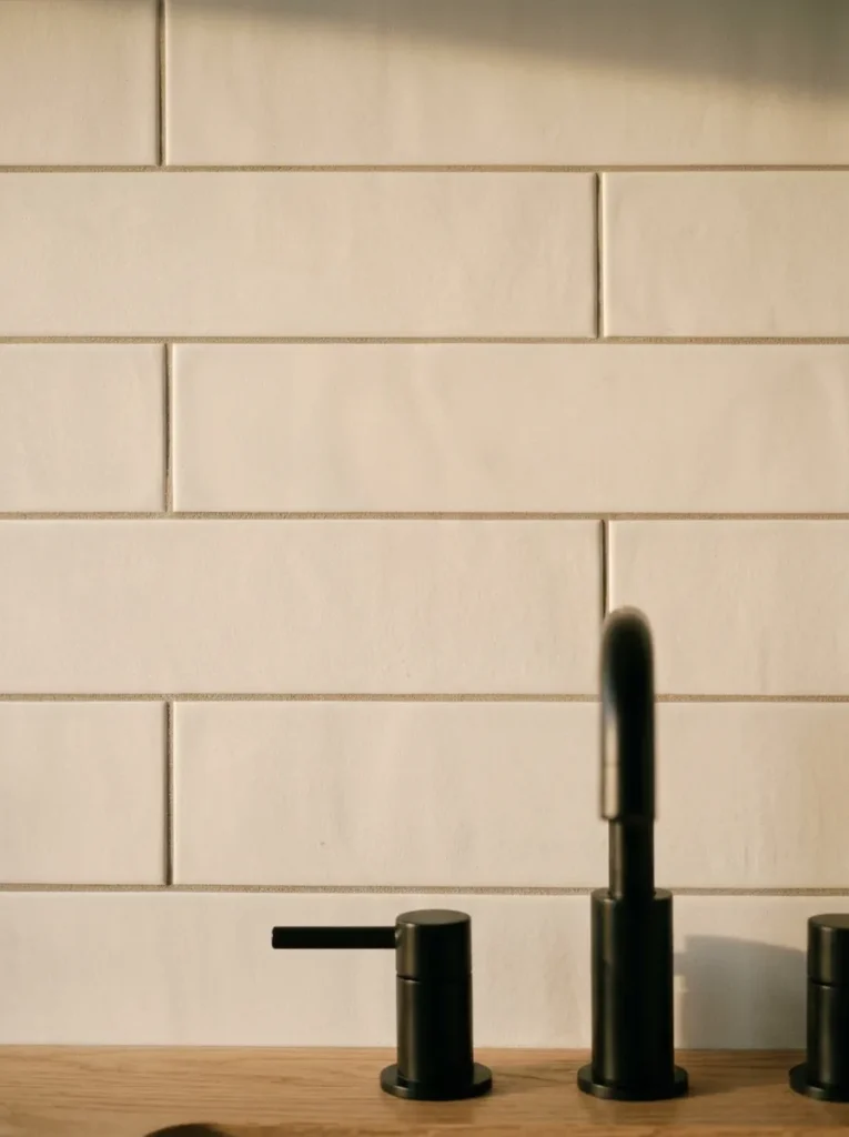 Matte white bathroom subway tile with warm-grey grout lines in raking afternoon light, matte black tap and pale oak vanity partially visible