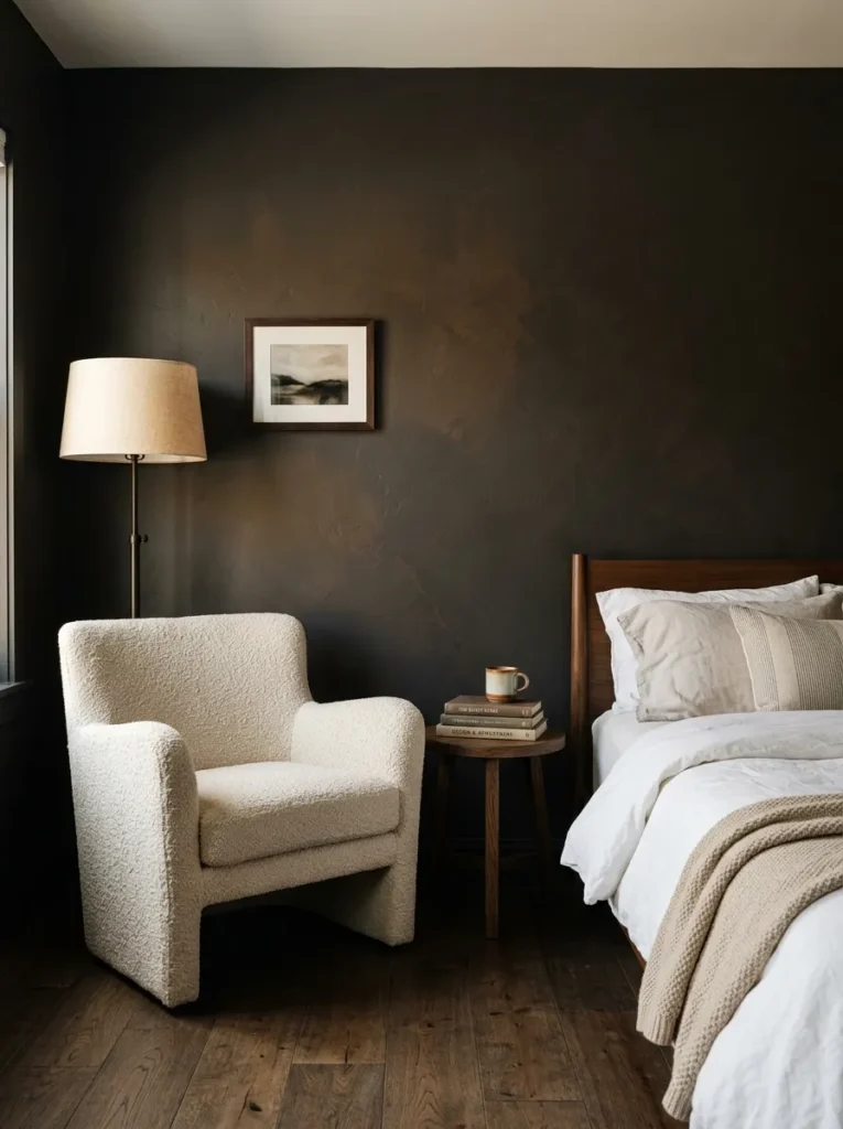 Bedroom painted in warm charcoal-black with brown undertones, a cream bouclé armchair, and soft afternoon light